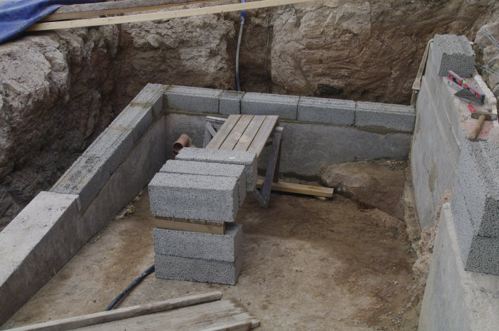 Grundarbetet för husbygge visar lecablocks, armering, betongsockel och grävd grop.