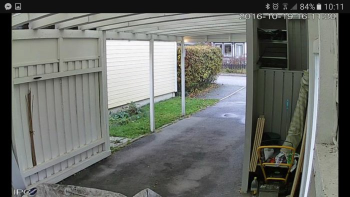 Övervakningskamera vy av en uppfart med carport, staket och en barnvagn synlig i hörnet.