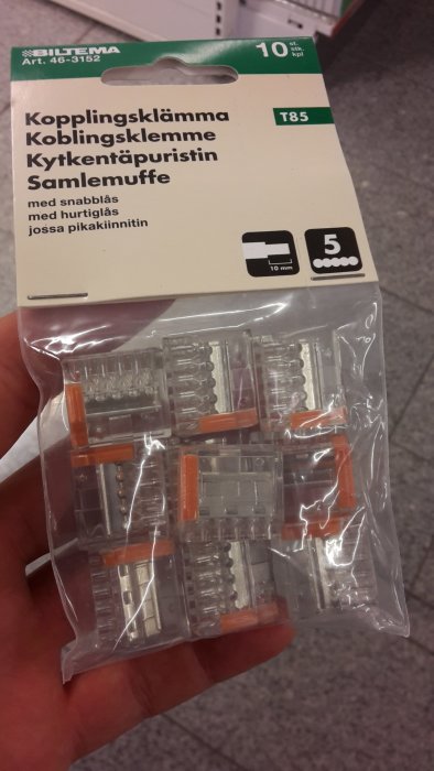 Hand håller en förpackning med transparenta kopplingsklämmor med orange spakar från Biltema.