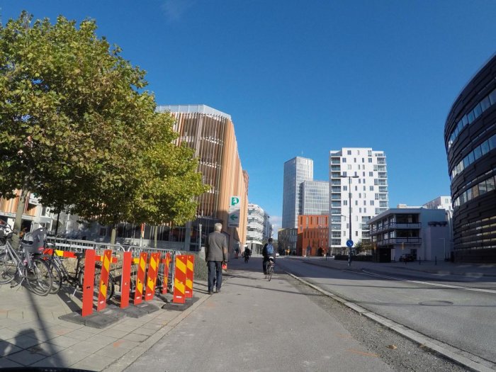 Klart blå himmel över en stadsgata med moderna byggnader, träd och personer.