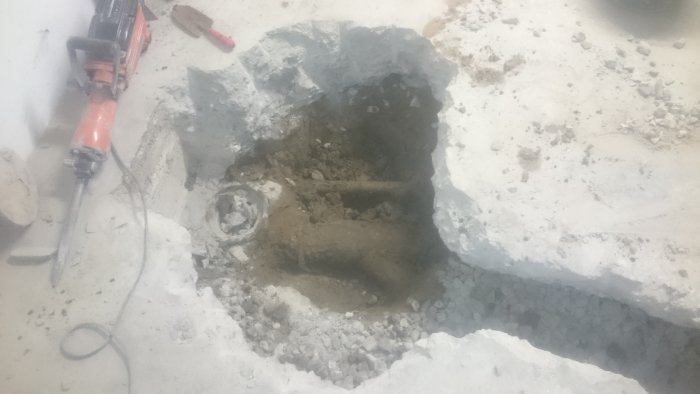 Grävt hål i betonggolv med synliga rör och en brytjärn, söker durkplåtslucka och backventil.