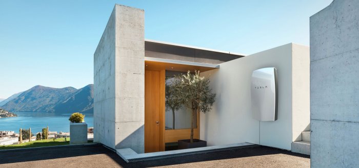 Modern villaentré med Tesla Powerwall tydligt monterad på väggen, berg och sjö i bakgrunden.