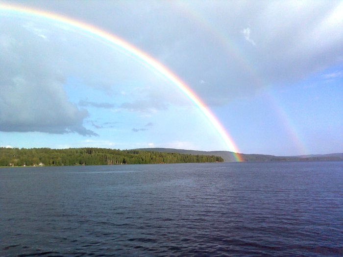 Dubbel regnbåge över en sjö med skogsbeklädda kuster i bakgrunden.
