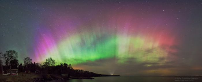 Panorama av ett intensivt norrsken i flera färger över Vättern med stjärnklar himmel.