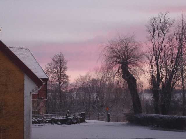 Vinterlandskap med snötäckt mark, träd, en röd byggnad och rosa himmel i Skåne.