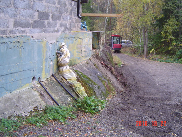 Delvis nedmonterad stödmur med gammal betong och verktyg framför, skog och grävmaskin i bakgrunden.