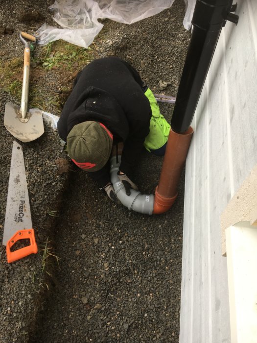 Person installerar avloppsrör vid husgrund med spade och såg i förgrunden.