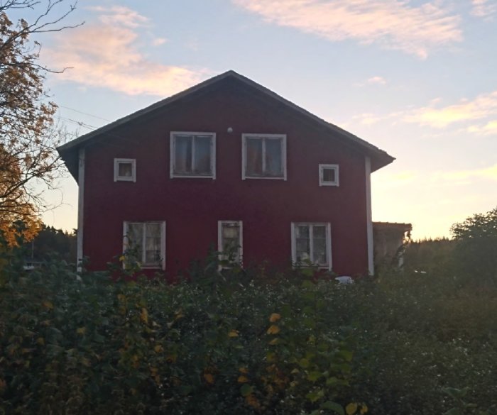 Rödfärgat hus med två våningar omgivet av grönska vid skymningen.