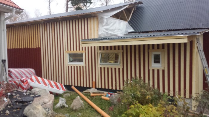 Pågående byggarbete på ett hus med röd och beige panel och ouppackade garagedörrar på marken.