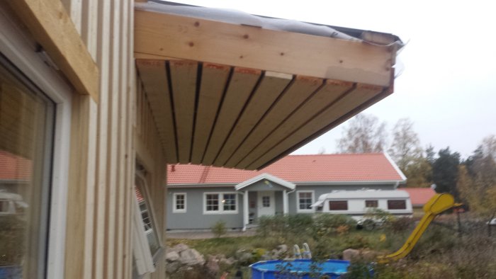 Taköverhäng på ett hus under konstruktion med utsikt över en annan villa och lekplats.