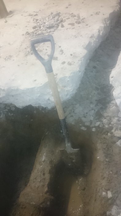 Spade stående i djupt, vattenfyllt hål för byggprojekt, med grus och bergkant synlig.