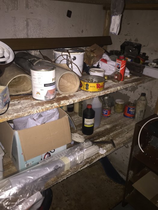 Källarförråd med fuktskador och diverse föremål som färgburkar och byggmaterial på en hylla.