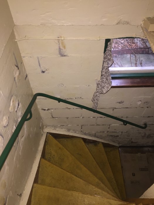 Trappa i källaren med fukt- och mögelskador på de vita väggarna och en bit tapet som hänger löst nära fönstret.
