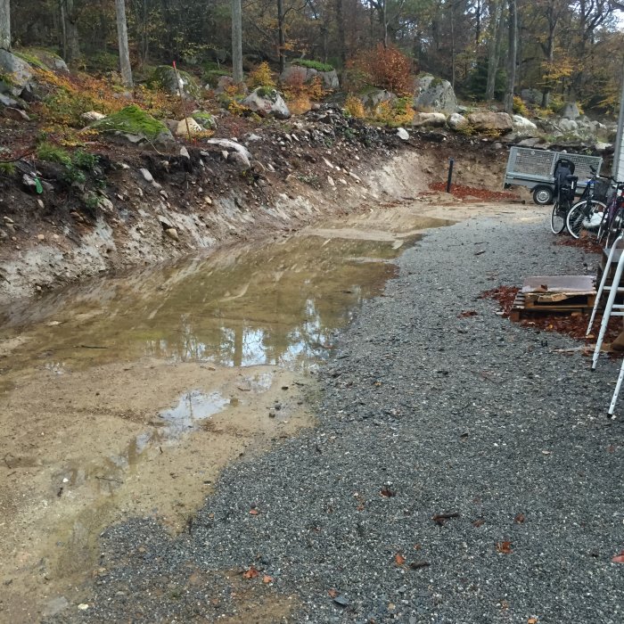 Stort vattenansamling längs nybyggd grund, dålig dränering, byggmaterial och cykel synliga.