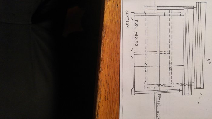 Teknisk ritning för byggprojekt med måttangivelser, delvis skymd av träkant.