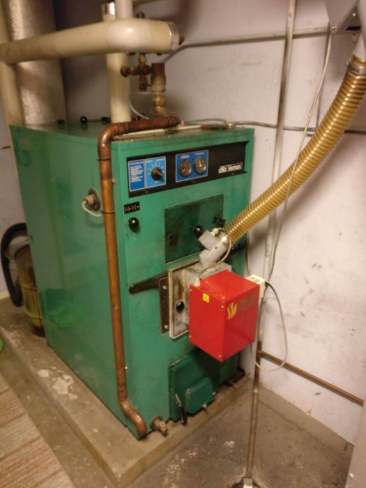 Gammal grön pannanläggning med kopplingar och röd eldningsautomatik i källare.