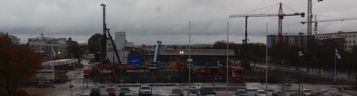 Byggarbetsplats med kranar, grävmaskiner och parkerade bilar under en molnig himmel vid lunchtid.