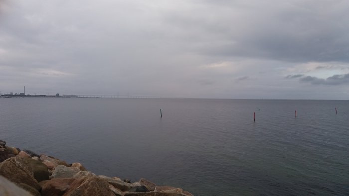 Molnig himmel över lugnt hav med stenig strandlinje och bro i bakgrunden.