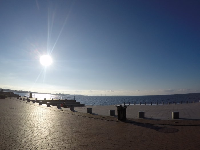 Solig höstdag vid en havspromenad med klarblå himmel och solen som reflekteras i vattnet.