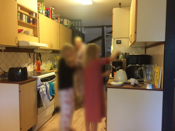 Gammalt kök med träskåp och vitvaror samt två suddiga personer.