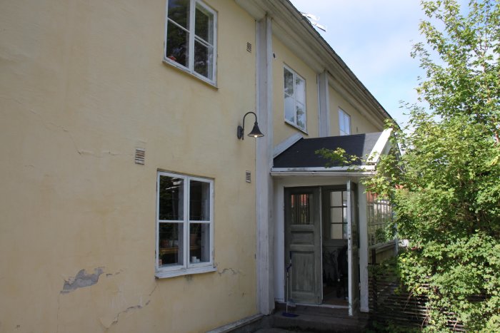 Gul kalkputsfasad på ett hus med krackelerande och flagnande områden vid fönster och hörn, i behov av renovering.