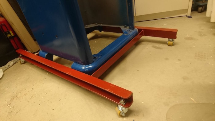 Monterade små hjul på en röd och blå sågmaskinsram i ett verkstadsutrymme, redo för mobilitet och stabilitetstestning.