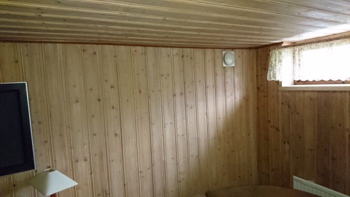 Vardagsrum med träpanel på väggar och i taket, ett fönster med gardiner till höger och en ventilationsöppning till vänster.