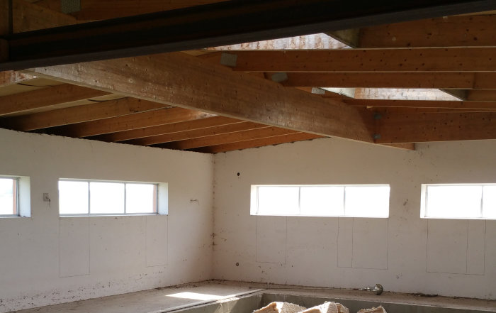Interior view of an unfinished room with exposed wooden beams and high windows, showing the progress of a ceiling renovation project.
