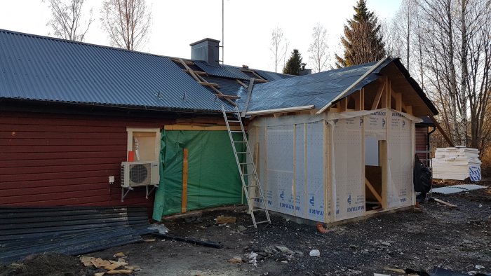 Nybyggnation av träkonstruktion med OSB-skivor vid rödlackerat hus, med byggmaterial och stege.