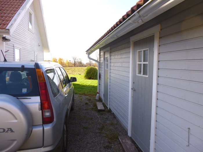 Gavel på vit träbyggnad med stupränna nära knuten, vid garage med vita dörrar och en parkerad bil.