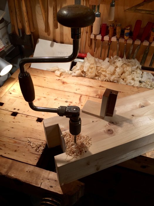 En gammaldags handdriven borrsväng i bruk på ett träarbetsbord med spån och verktyg i bakgrunden.