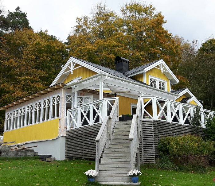 Gult sekelskifteshus i renovering med delvis nybyggd glasveranda och borttagen ytterpanel, omgivet av grönska.