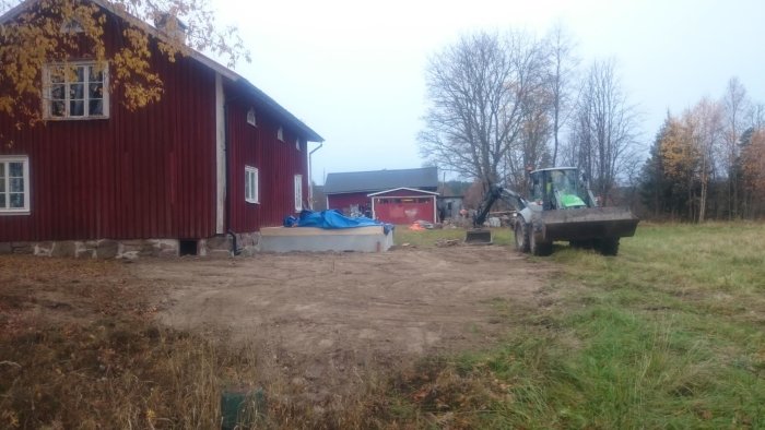 Grävtraktor som jämnar marken runt en tillbyggnad av ett rödmålat hus, med tidigare lerhögar borttagna.