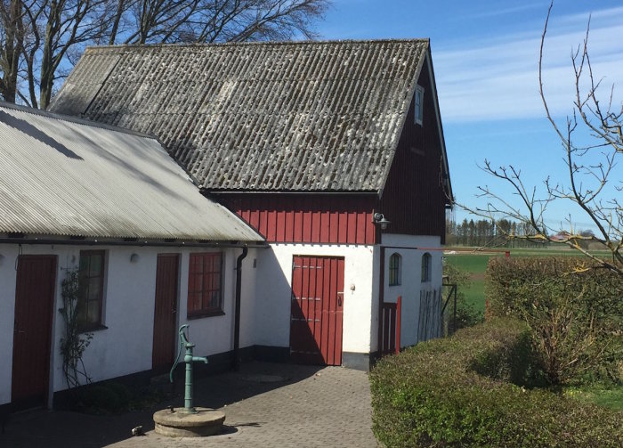 Gammal ladugård med kalkputsat tegel och eternittak före ombyggnad till hemmakontor, synlig handpump i förgrunden.