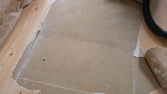 Uppriven gammal plastmatta som avslöjar skadat golvspån med en synlig kant.