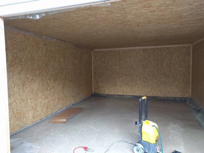 Garage interiör före målning med OSB-väggar och tak, och en målarspruta på golvet.