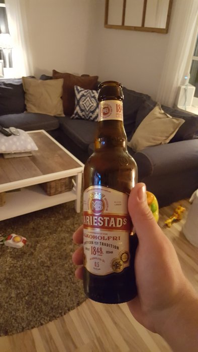 Hand håller Mariestads alkoholfri öl i vardagsrum med soffa och bord i bakgrunden.