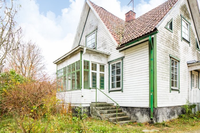 Ett slitet vitmålat hus med gröna fönsteromfattningar och en veranda, omgivet av vildvuxen vegetation.