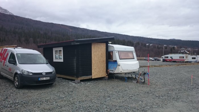 Nybyggd liten stuga med oskyddad fasad och en husvagn på en grusplan med bakgrund av skog och berg.