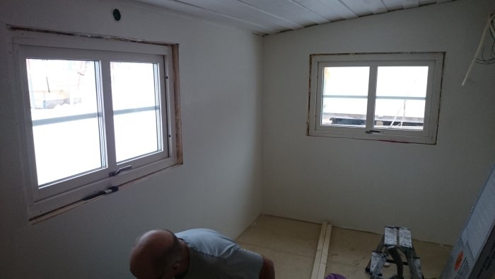 Interiör av ett rum under renovering med omonterade dörrkarmar runt två fönster och en person som arbetar.