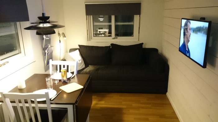 Vardagsrum med soffa, matbord och TV på väggen.