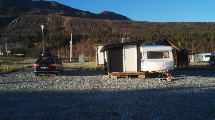 Husvagn med förtält parkerad på grusplats med en bil framför mot bergig bakgrund i dagsljus.