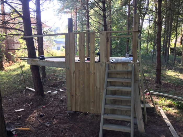 Pågående konstruktion av ett träfort i skogsmiljö med stegar, verktyg och osorterat byggmaterial synligt.