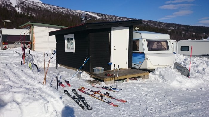 Husvagn och modulär utebyggnad på campingplats i snölandskap med skidor och stavar utanför.