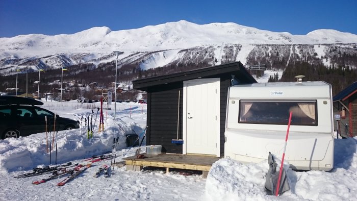 Husvagn parkerad vid snöig campingplats med skidor, skidstav, husbil och berg i bakgrunden.
