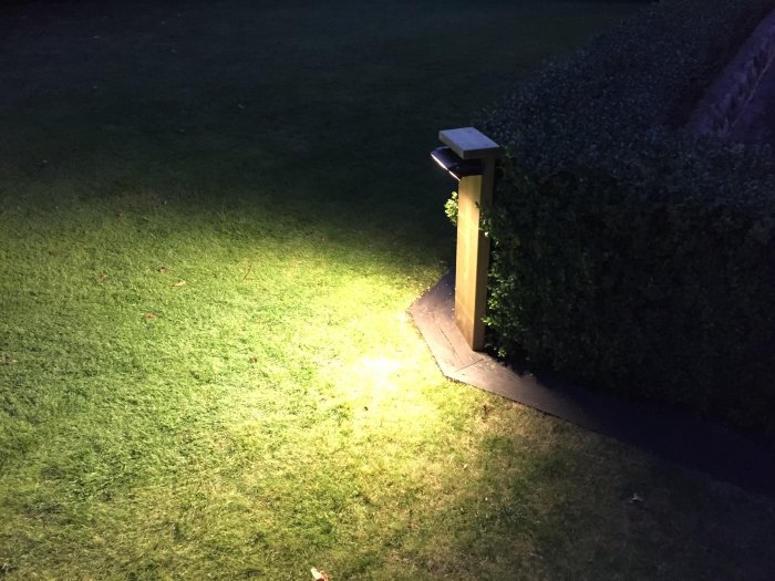 En svart utomhuslampa monterad på tryckt virke vid en häck lyser upp en gräsmatta på kvällen.