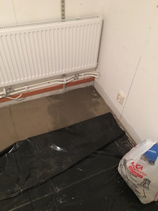 Renoveringsprojekt visande en hörna med radiator, blottlagd betongplatta och svart plastfolie på golvet.