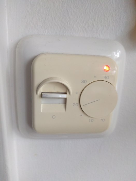 Gammal beige termostat för golvvärme på vägg med röd lampa tänd och vred inställt på cirka 20 grader Celsius.