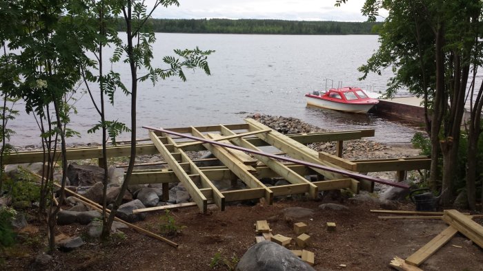 Byggprojekt vid vattnet med träreglar och fundament, en båt i bakgrunden.