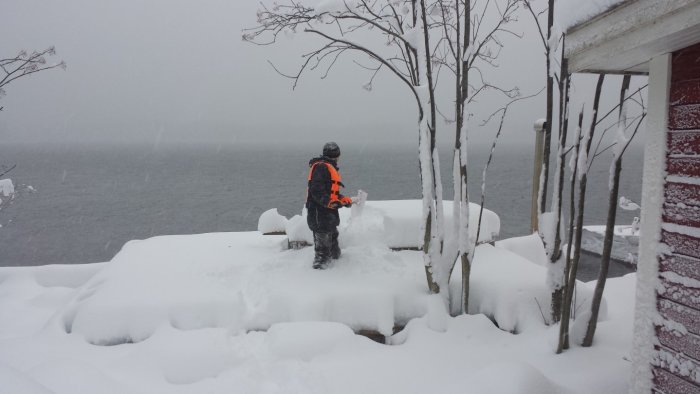 Person i reflexväst schaktar snö nära vatten vid kant av snötäckt terrass under snöfall.